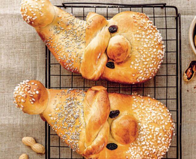 Dwie bułki Drożdżowe Mikołaje w kształcie twarzy Świętego Mikołaja, posypane nasionami i rodzynkami, ułożone na podstawce do studzenia.