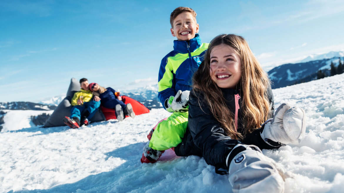 Zabawa na śniegu nawet bez czapek © SalzburgerLand Tourismus, Markus Berger Dwoje uśmiechniętych dzieci bawi się na śniegu, ciesząc się nartami w wiosennym słońcu, podczas gdy dorośli relaksują się na workach z fasolą, góry za nimi. (Powiększony widok)