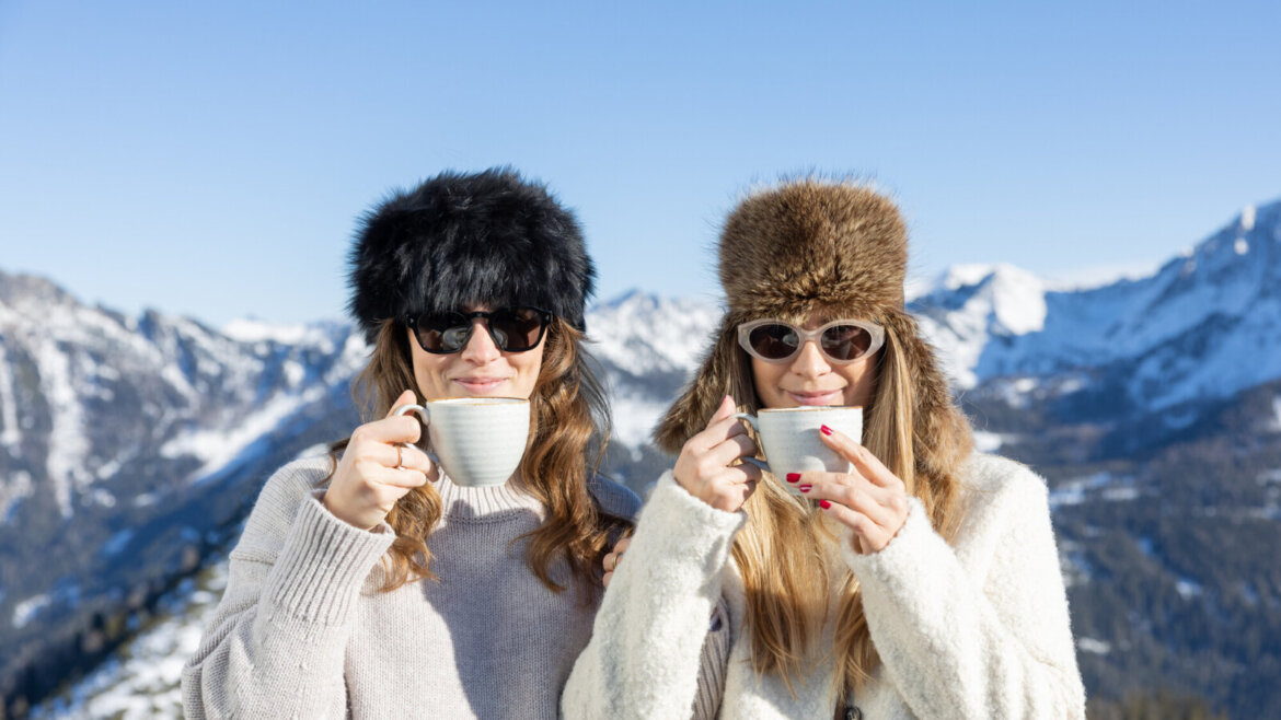 Ciesz się zimą w promieniach wiosennego słońca © SalzburgerLand Tourismus, Hansi Heckmair Dwie kobiety w futrzanych czapkach cieszą się kubkami, chłonąc narty w wiosennym słońcu ze śnieżnymi szczytami za sobą. (Powiększony widok)