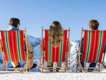 rs6713_salzburg_chill_out1341-lpr-1920×658.jpg Trzy osoby na leżakach w paski cieszą się narty w wiosennym słońcu, z widokiem na śnieżne góry i czyste, błękitne niebo.
