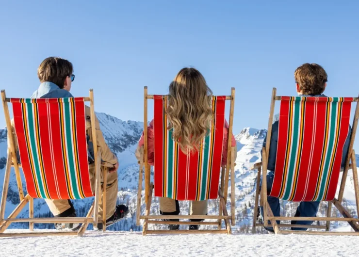 Trzy osoby na leżakach w paski cieszą się narty w wiosennym słońcu, z widokiem na śnieżne góry i czyste, błękitne niebo.
