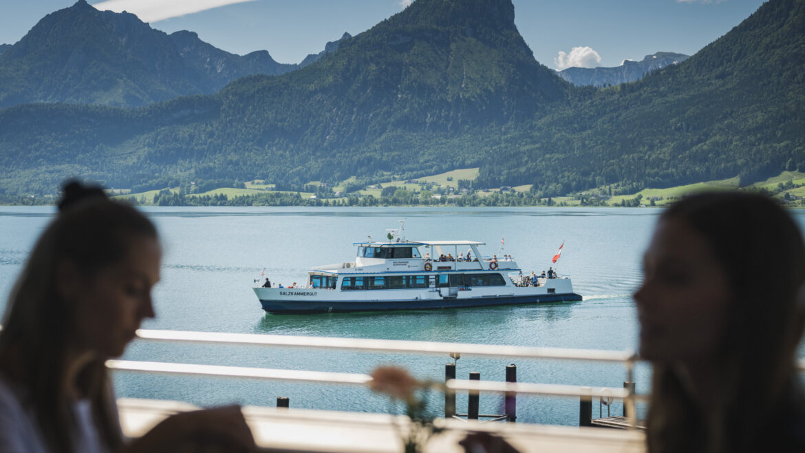 Kawa i ciasto na tarasie restauracji przy stacji kolejki na Schafberg, z widokiem na jezioro Wolfgangsee © Salzburg AG Tourismus GmbH Dwie osoby siedzą nad jeziorem w Salzburgu Salzkammergut, z łodzią i górami pod błękitnym niebem. (Powiększony widok)