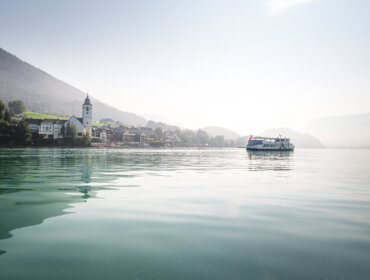 Łódź pływa po spokojnym jeziorze Salzkammergut w pobliżu wiejskiego kościoła, z górami w tle.