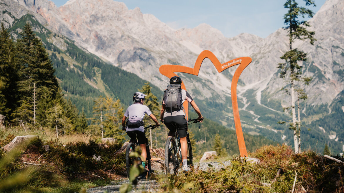 Dwóch rowerzystów zjeżdża z góry na rowerach górskich wzdłuż szlaku z górami i dużą pomarańczową ramą za nimi.