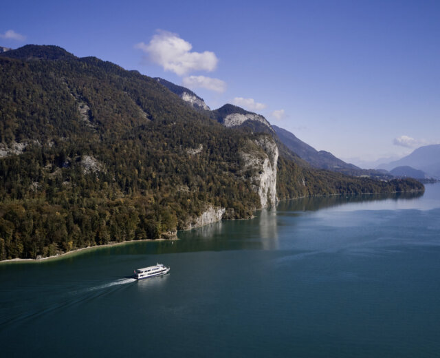 Łódź płynie po jeziorze Wolfgangsee, otoczona zalesionymi górami pod czystym, błękitnym niebem. Rejsy statkiem.