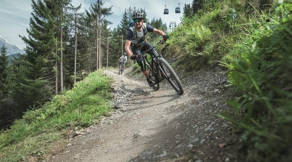 Dwóch rowerzystów górskich ściga się w słoneczny dzień po krętej, polnej ścieżce prowadzącej przez zalesione wzgórze.