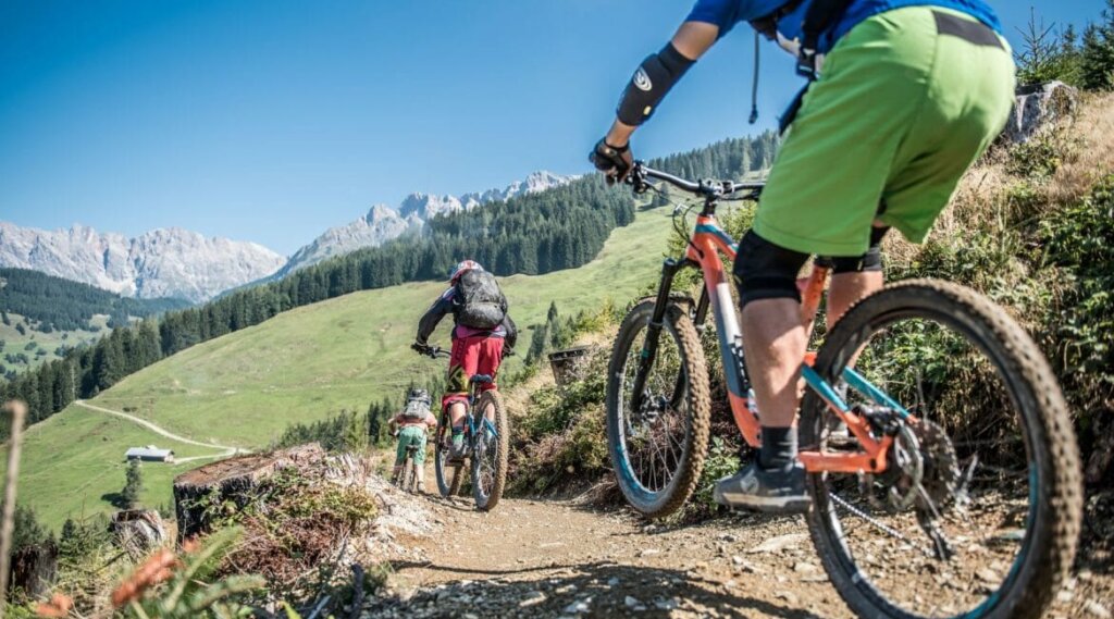 Trzech rowerzystów górskich ściga się w dół po polnej ścieżce przez trawiaste wzgórza z górami w tle.