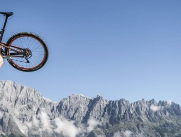 hochkonig-sommer-biken-1920×658 Osoba w niebieskiej koszuli podnosi rower górski ze skalistymi górami, pokazując turystykę rowerową w Hochkönig.