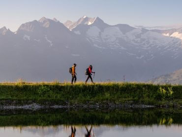 hohe-tauern-panorama-trail-etappe-03tvb-wald-koenigsleiten-1920×1067 Para wędrowców idzie brzegiem górskiego jeziora, mając w tle potężne masywy górskie Wysokich Taurów.