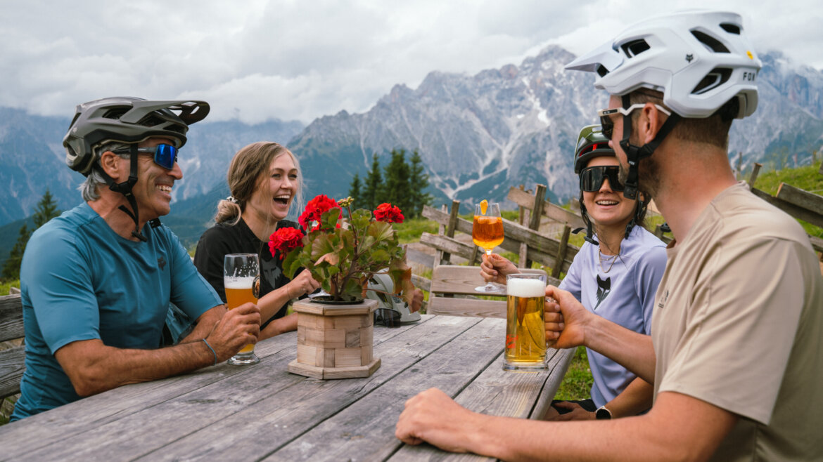 Czterech rowerzystów w kaskach pije drinki i śmieje się, a ich górskie Hochkönigi zaparkowane są nieopodal.