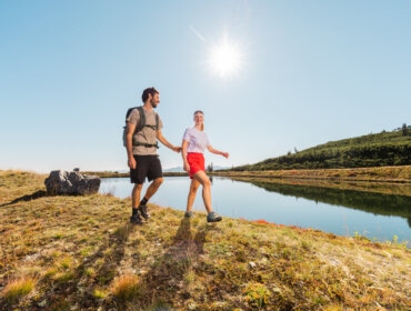 ©Salzburger Lungau (4) Mężczyzna i kobieta wędrują w słoneczny dzień obok spokojnego jeziora Być.Sobą latem w Lungau.