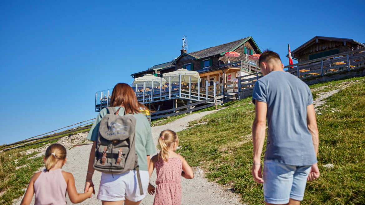 Czteroosobowa rodzina idzie żwirową ścieżką w kierunku schroniska górskiego w pobliżu kolejki zębatej na Schafberg. (Powiększony widok)
