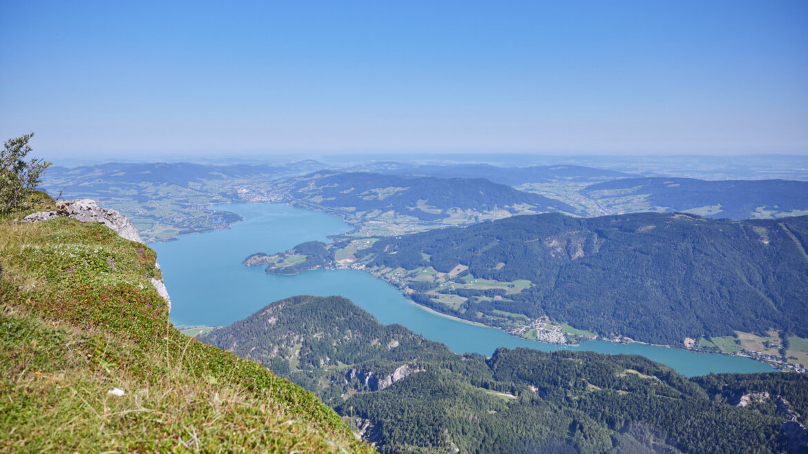 Widok z lotu ptaka na błękitne jezioro, zielone wzgórza i kolej zębatą na górę Schafberg wijącą się przez góry. (Powiększony widok)
