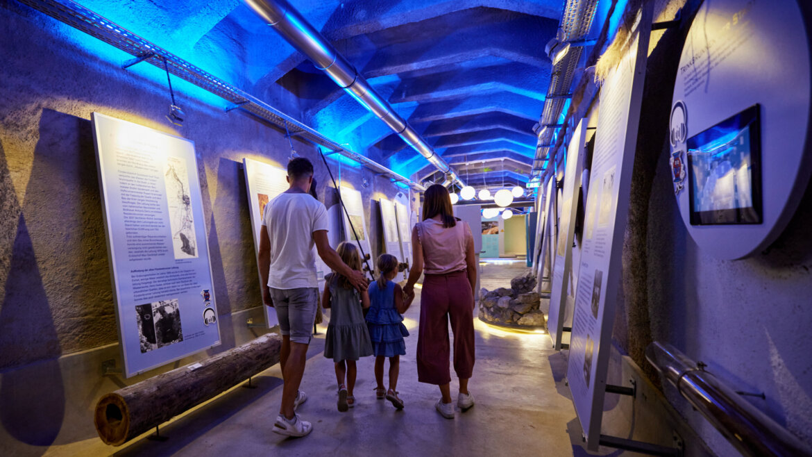 Rodzina zwiedza oświetlone na niebiesko muzeum wody WasserSpiegel, z panelami wystawowymi i informacjami na ścianach. (Powiększony widok)