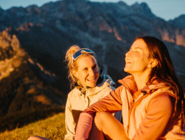 Dwie kobiety cieszą się turystyką pieszą w regionie Hochönig, śmiejąc się na trawie z górami o zachodzie słońca.