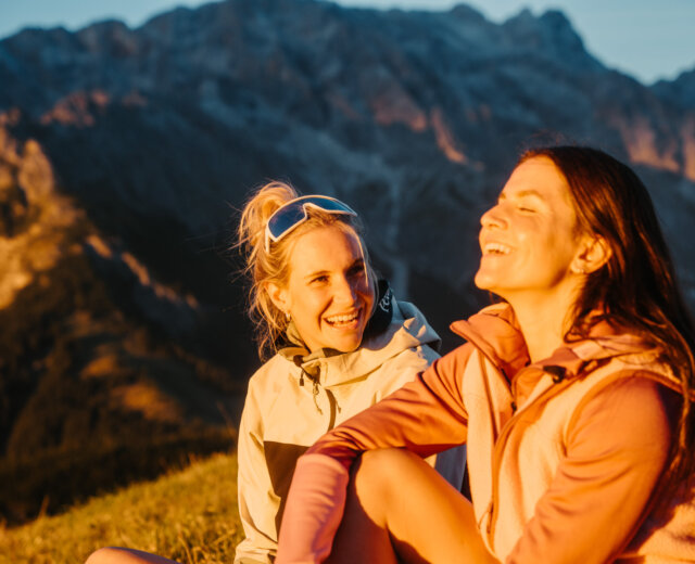 Dwie kobiety cieszą się turystyką pieszą w regionie Hochönig, śmiejąc się na trawie z górami o zachodzie słońca.