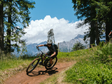 Rowerzysta górski w kasku zjeżdża w dół po polnej ścieżce otoczonej drzewami i górami.