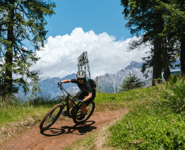 Rowerzysta górski w kasku zjeżdża w dół po polnej ścieżce otoczonej drzewami i górami.