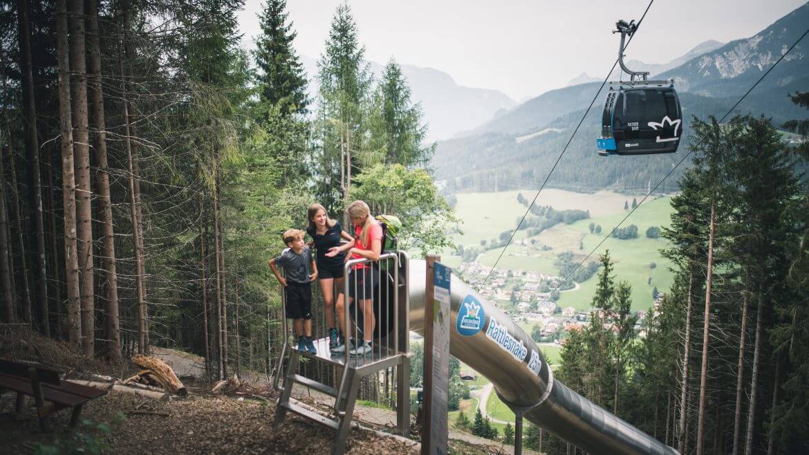 Trójka dzieci przed wejściem do zjeżdżalni, jednej z 5 zjeżdżalni z góry Natrun w dolinę. Ponad ich głowami mknie gondola kolejki górskiej.