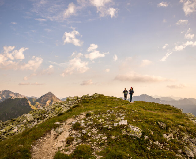 Dwóch turystów wędruje o wschodzie słońca na szczyt Speiereck w salzburskim regionie Lungau. Mogą podziwiać imponujący otaczający krajobraz górski i oglądać wschód słońca.