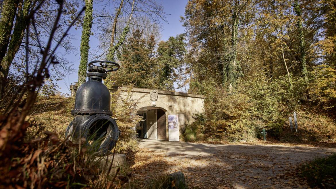 Kamienne wejście do budynku w pobliżu jesiennego lasu, z dużym metalowym zaworem - częścią muzeum wody WasserSpiegel. (Powiększony widok)