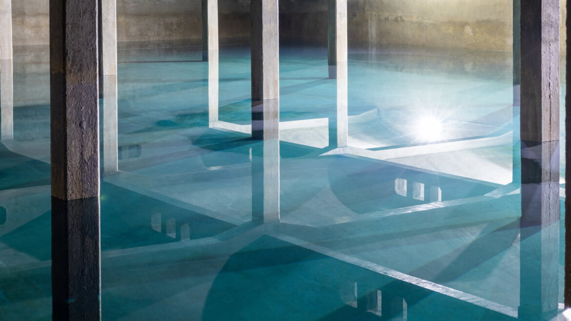 Betonowe filary odbijają się w czystej, błękitnej wodzie wewnątrz rozległego podziemnego zbiornika Muzeum Hydrotechniki. (Powiększony widok)