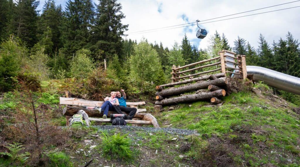 Para wędrowców odpoczywa na drewianej ławce na skraju górskiego lasu. Ponad ich głowami mknie wagonik kolejki linowej