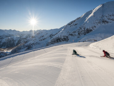 Dwóch narciarzy zjeżdża ze świeżo przygotowanego zaśnieżonego stoku w jasnym słońcu w górskim krajobrazie.