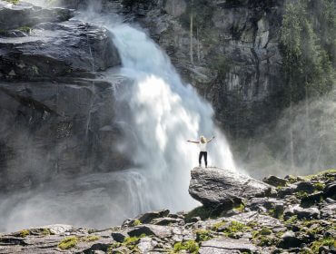 Na głazie u stóp wodospadu w Krimml stoi kobieta, która podnosi ramiona w górź i wdycha leczniczą mgiełkę wodną.