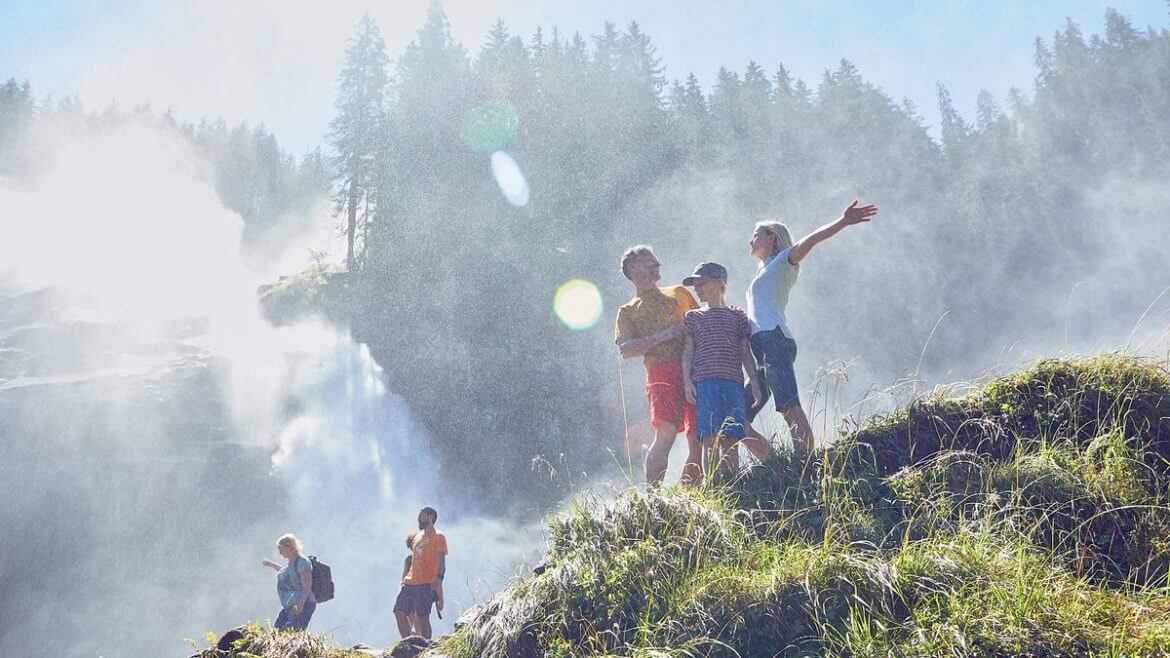 Turyści u stóp wodospadu w Krimml pełną piersią wdychają otaczającą ich mgiełkę wodną. W tle dolna kaskada największego wodospadu w Europie.