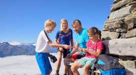 Rodzice z dwójką nastolatków zdobyli jeden ze szczytów w Wysokich Taurach. Dzieci próbują rozerwać kiełbaski. Błękitne niebo nad głowami, gęste chmury poniżej.