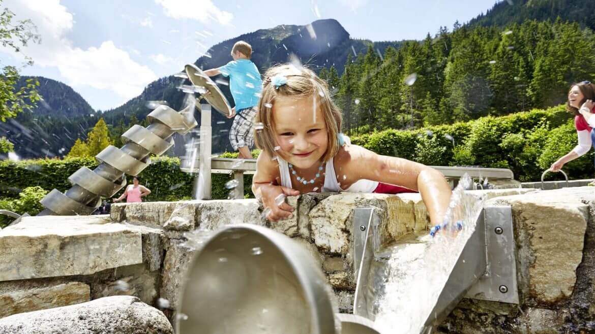 W słoneczny dzień w familijnym parku rozrywkowo-edukacyjnym WasserWunderWelt mama z córką i synem bawią się radośnie wodą przy przy pompach i rynnach. W tle szczyty Wysokich Taurów.
