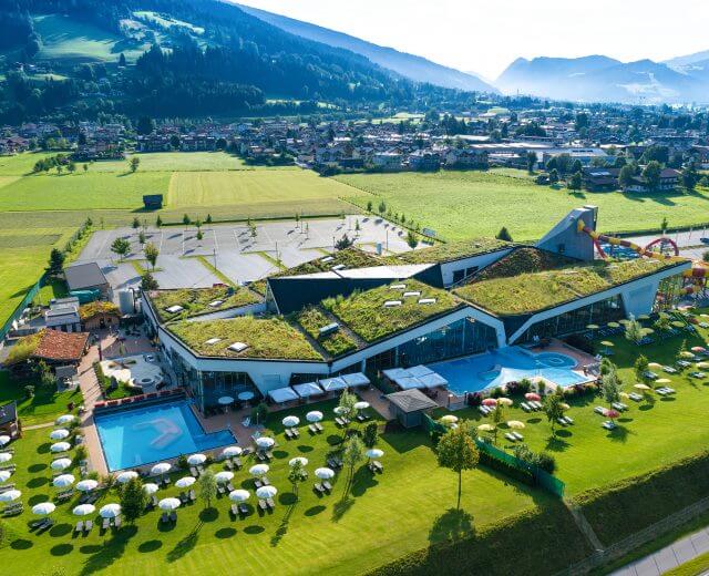 Widok basenów termalnych Erlebnis Therme Amadé w Altenmarkt-Zauchense z lotu ptaka. Zielone łaki z leżakami i baseny z błękitną wodą otaczają łańcuchy górskie.