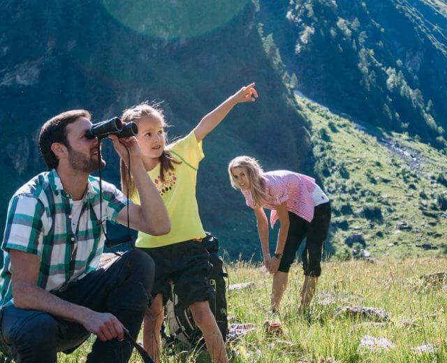 Górska polana, a na niej tato z córką obserwują przez lornetkę prawdopodobnie lecącego po niebie orła, w tle mama podwija spodnie.