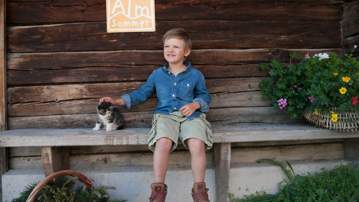 Chłopiec na ławce przed drewanianą bacówką głaszcze siedzącego obok kota. Z jego drugiej strony olbrzymi wiklinowy kosz z kwiatami. Nad nim tablica z certyfikatem "Lata na Halach".
