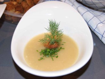 Zupa - musjabłkowy z chrzanem, podna na białej miseczce, przybrana posiekanym szczypiorkiem i gałączkami koperku.