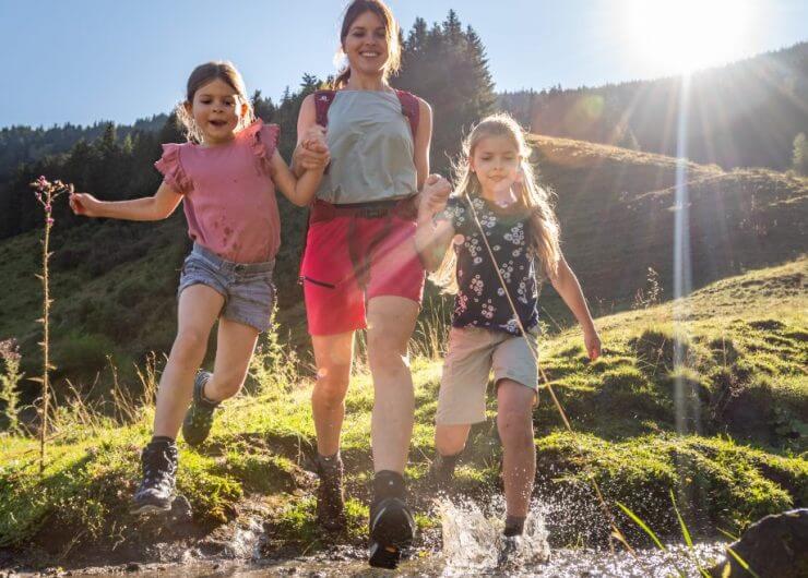 Po alpejskiej łące biegnie mama z dwiema dziewczynkami. Przeskakują przez górski potok. Dziewczynki trzymają mamę za ręce. Wszystkie trzy są radosne i roześmiane.