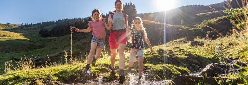 Po alpejskiej łące biegnie mama z dwiema dziewczynkami. Przeskakują przez górski potok. Dziewczynki trzymają mamę za ręce. Wszystkie trzy są radosne i roześmiane.