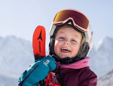 Uśmiechnięte dziecko w sprzęcie narciarskim trzyma narty na ośle łączki Dachstein West, gotowe do nauki jazdy na nartach.