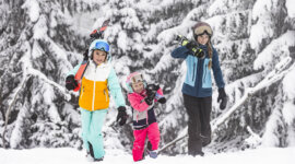 Trzy osoby w kolorowych zimowych ubraniach niosą deski snowboardowe i narty - idealne ferie zimowe z dziećmi w zaśnieżonym lesie.