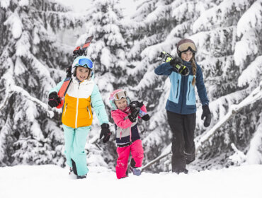 Familienskigebiet Dachstein West (c) Dachstein Tourismus AG, MirjaGeh (1) Trzy osoby w kolorowych zimowych ubraniach niosą deski snowboardowe i narty - idealne ferie zimowe z dziećmi w zaśnieżonym lesie.