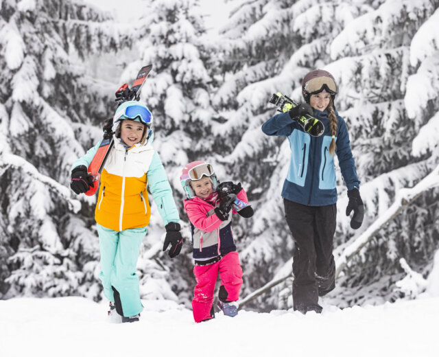 Trzy osoby w kolorowych zimowych ubraniach niosą deski snowboardowe i narty - idealne ferie zimowe z dziećmi w zaśnieżonym lesie.