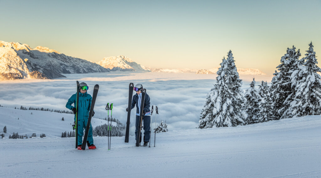Dwóch narciarzy na zaśnieżonym stoku w ośrodku Zima w Dachstein West, ponad chmurami i wśród pokrytych śniegiem drzew.