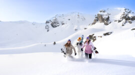 Trzy osoby w zimowych ubraniach idą przez głęboki śnieg - Jesteśmy pokoleniem zimy - niosąc deski snowboardowe.