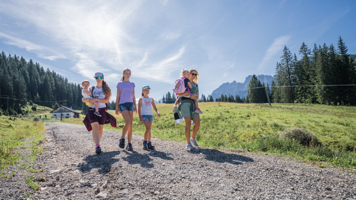 Dwie młode mamy z czwórką dzieci wędrują szutrowym szlakiem, niosąc najmłodsze na ręku, w tle las i góry Dachstein West.
