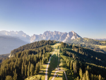 Widok z lotu ptaka na krętą górską drogę w pobliżu Dachstein West, otoczoną zielonymi lasami i skalistymi szczytami pod czystym niebem.