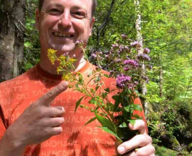 Mężczyzna w pomarańczowej koszuli uśmiecha się i trzyma krwawnik i dzikie oregano w bujnym, zielonym lesie.