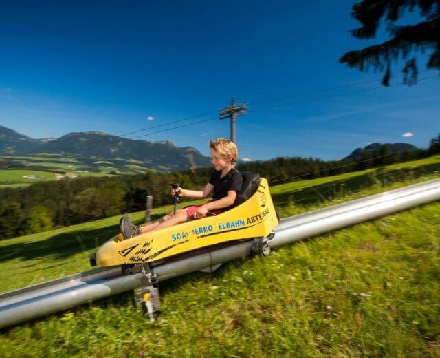 sommerrodeln-karkogel_abten-1920×1067-1-1170×658 Chłopiec w żółtych saneczka zjeżdża w dół po letnim torze saneczkowym na Karkogel w Abtenau.