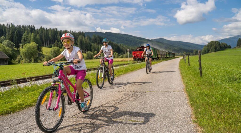Trzyosobowa rodzina na rekreacyjnej wycieczce rowerowej w dolinie Salzburskiego Lungau. W tle widoczna wąskotorowa kolejka Taurachbahn.