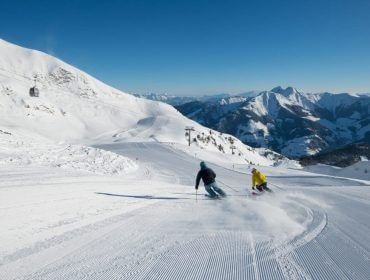 Dwóch narciarzy na nartach przyjaznych dla środowiska zjeżdża po zaśnieżonym stoku z górami i wyciągiem narciarskim za nimi.